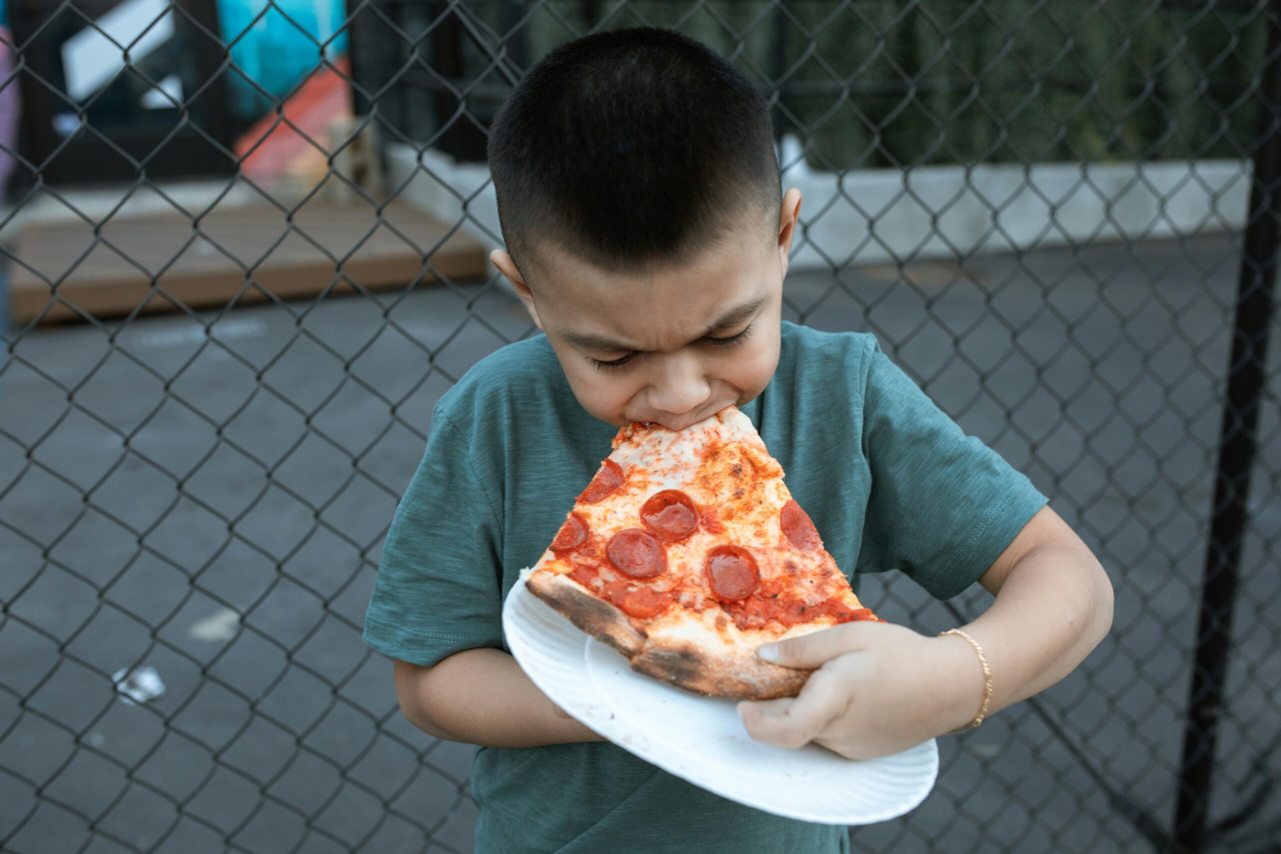 A child in a green shirt eagerly bites into a large pepperoni pizza slice outdoors.